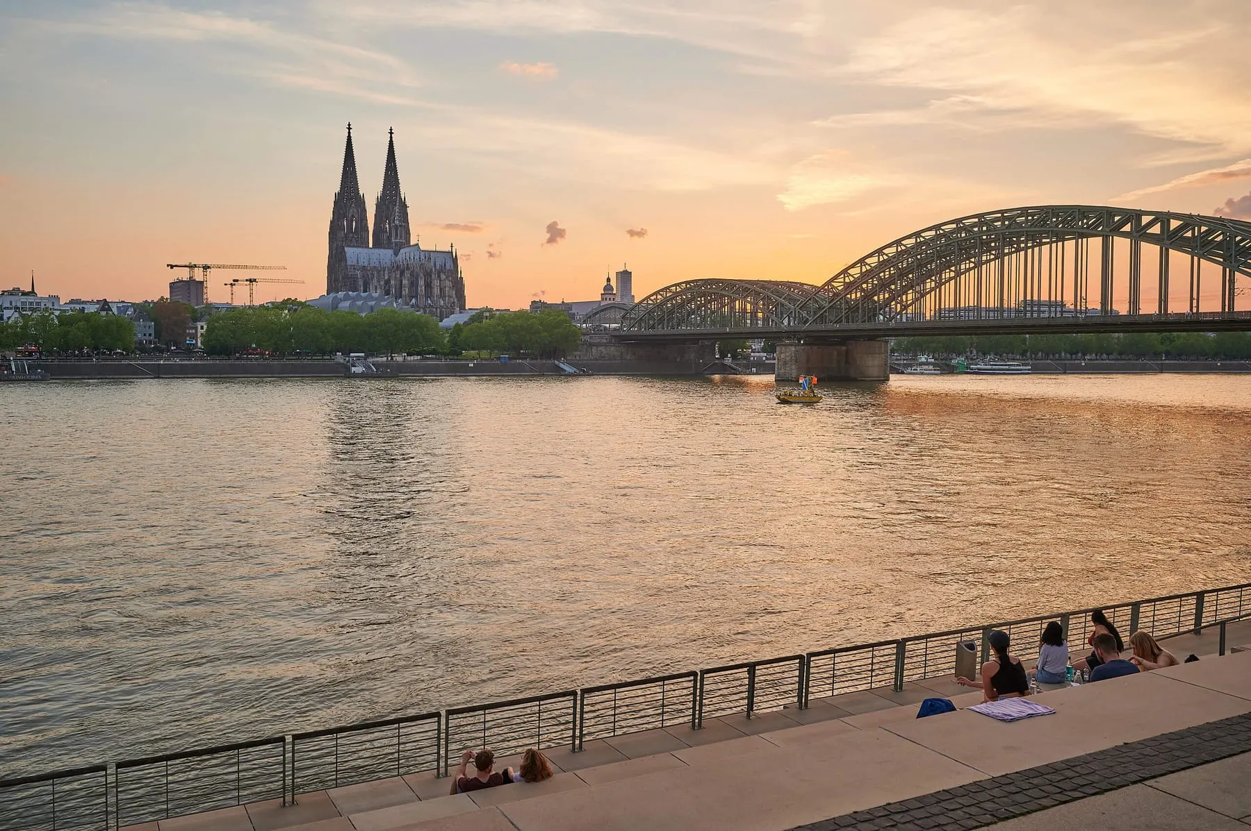 Rheinufer in Köln-Deutz im Abendlicht: Blick über den Rhein zum Kölner Dom und zur Hohenzollernbrücke, Menschen sitzen auf der Freitreppe am Wasser